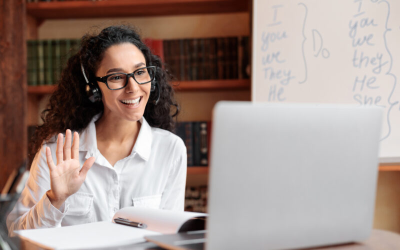 Distance Education. Positive lady wearing glasses and wireless headest at virtual meeting, sitting at desk, having video call on laptop, waving to webcam. Woman studying or teaching online at home